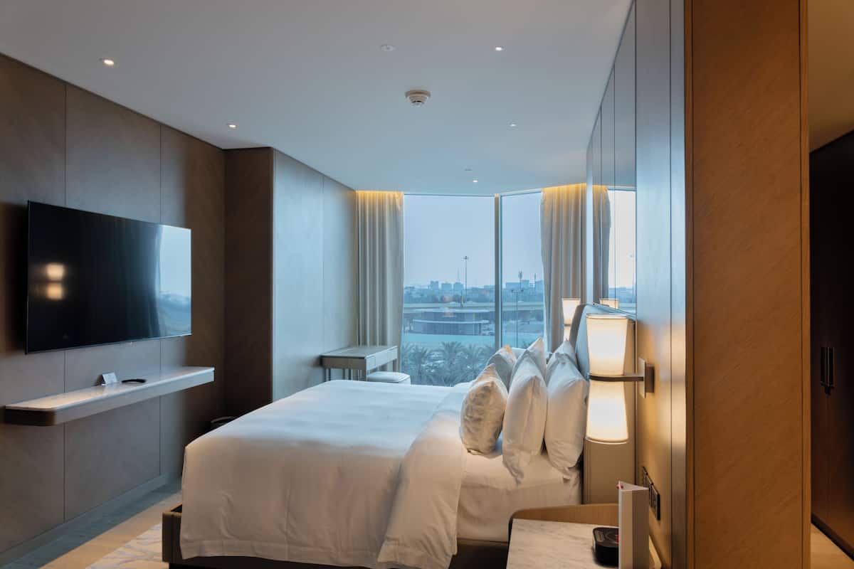 A modern hotel room with a large bed, a flat-screen TV mounted on the wall, a bedside table, and a view of the city through large windows.