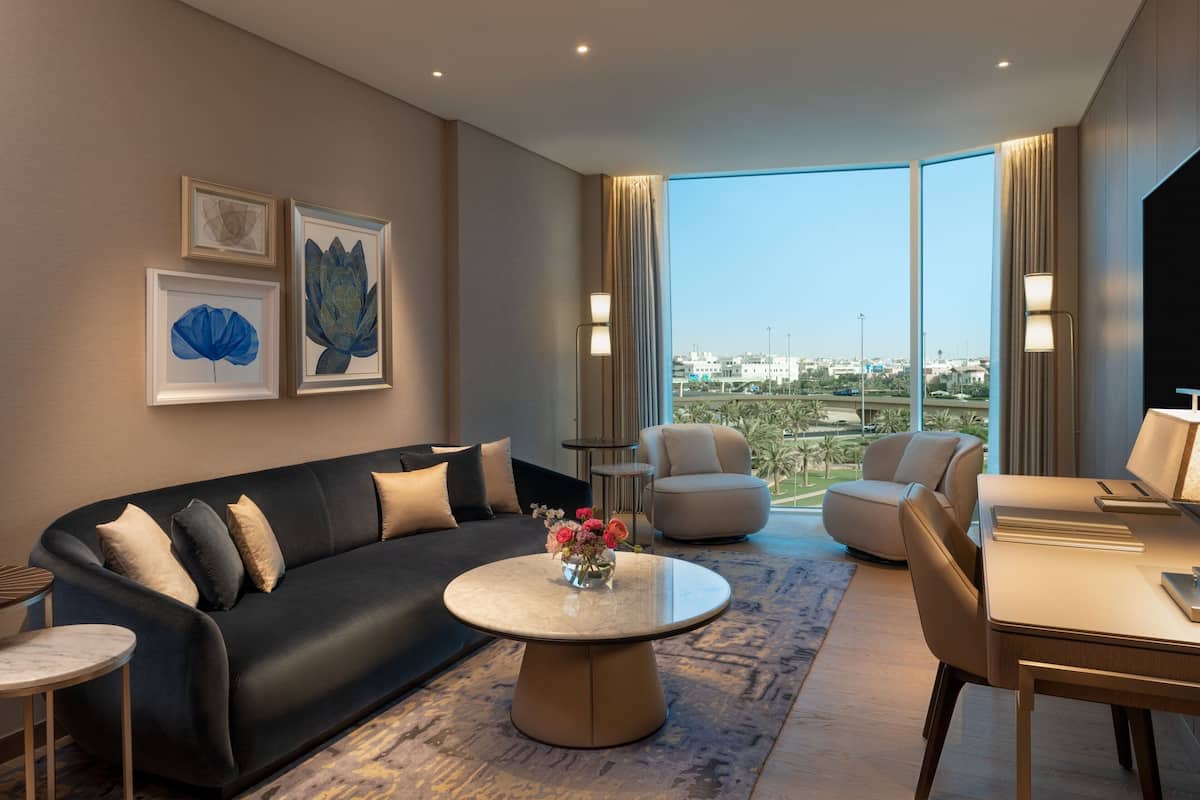 A modern hotel room with a black leather sofa, a round coffee table with a vase of flowers, a dining table with chairs, and large windows offering a city view.
