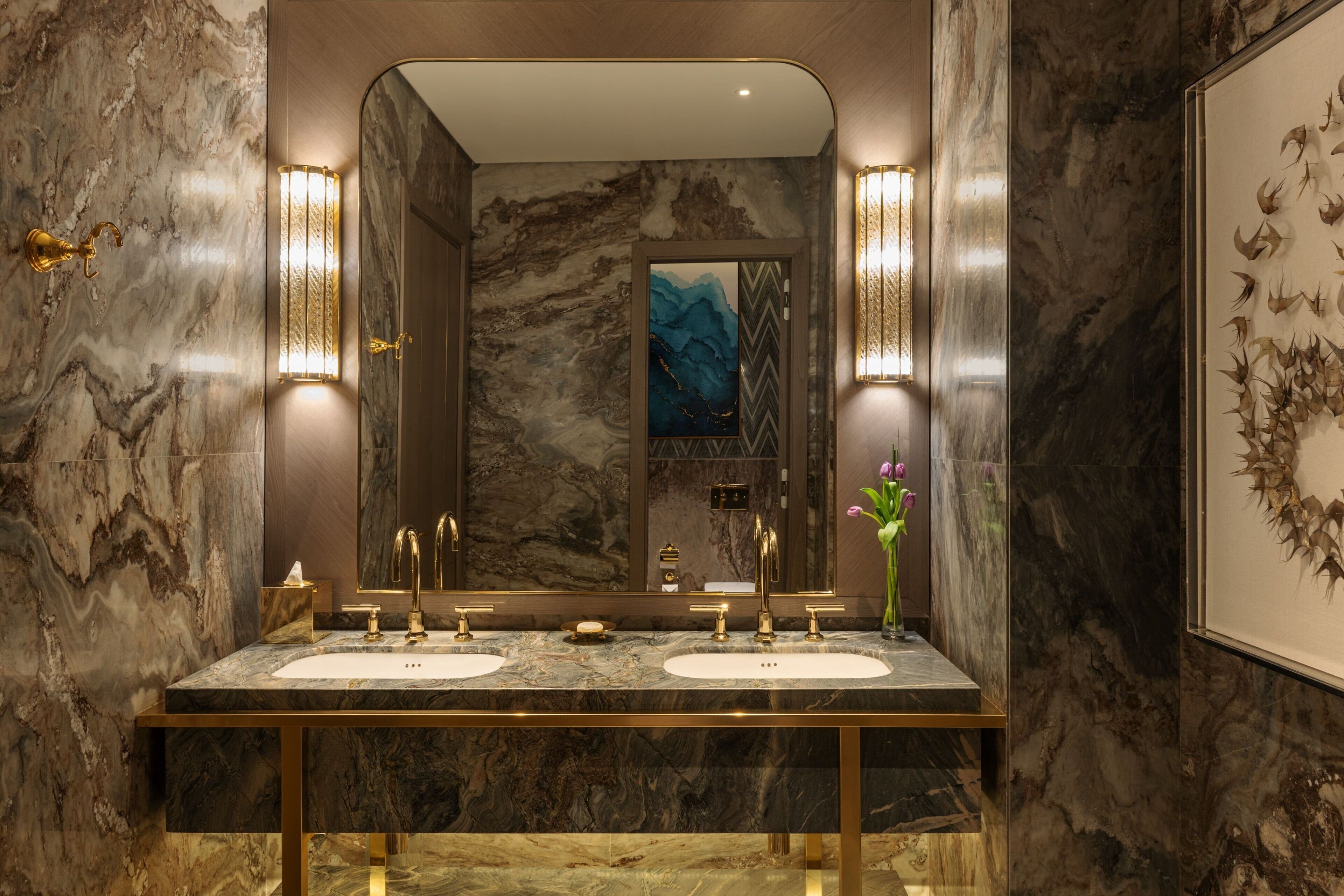 A bathroom with two sinks, marble walls, gold fixtures, and wall-mounted lights.