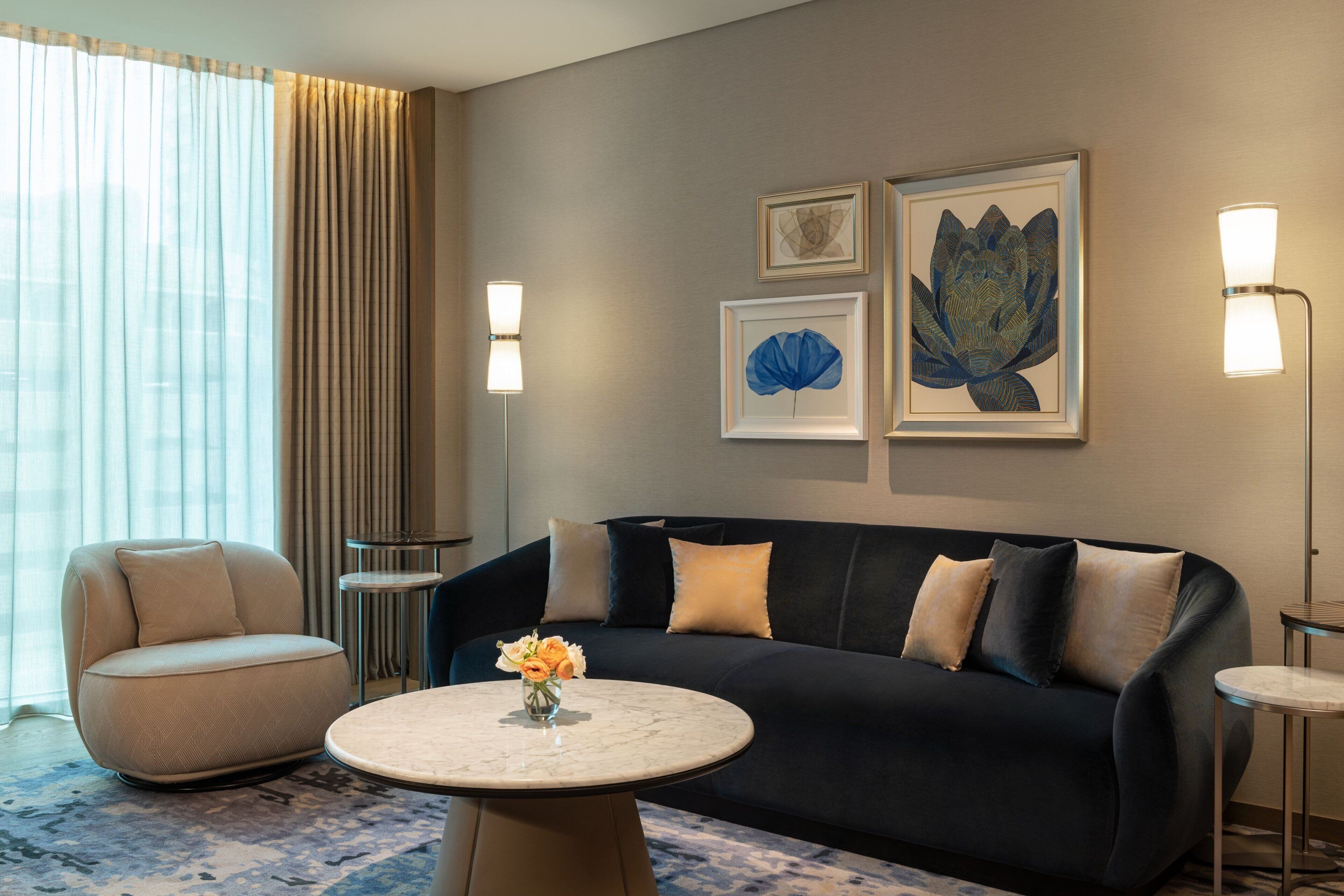 A modern living room with a dark sofa, a round coffee table, and a beige armchair. There are two framed artworks on the wall and a floor lamp.