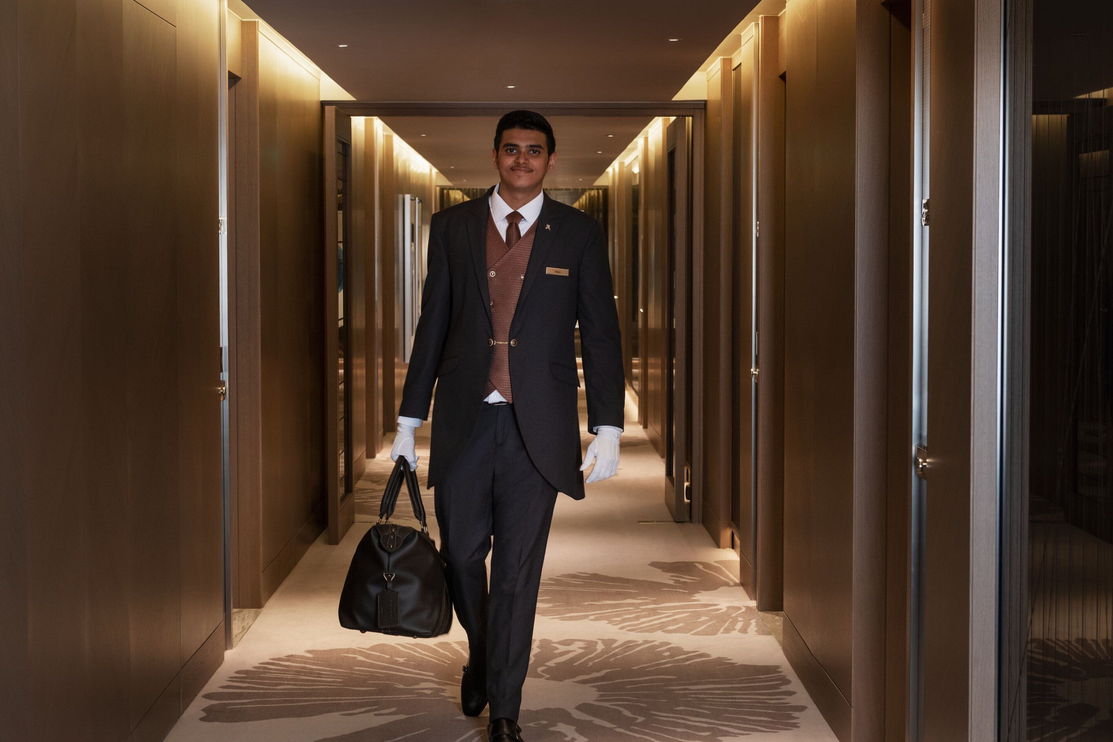 A man in a dark suit and tie walking down a hallway with wooden doors and a patterned carpet.