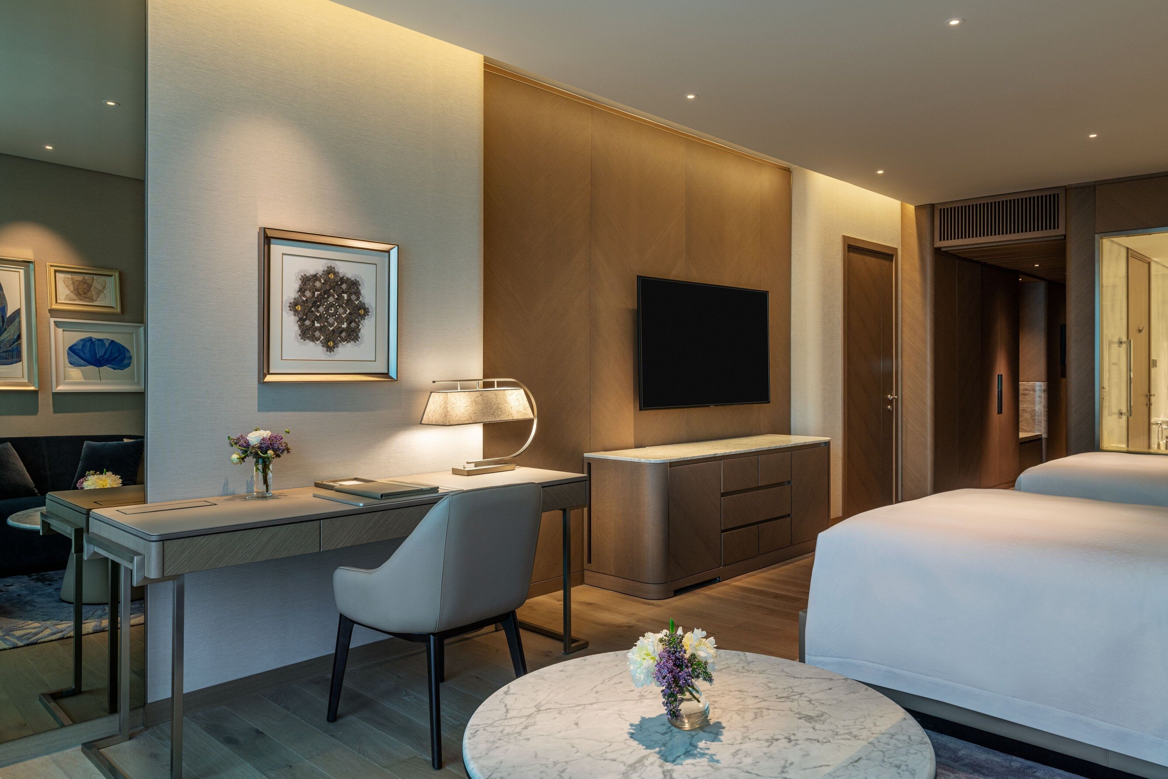 A modern hotel room with a desk, chair, round table with flowers, a TV, and two beds.