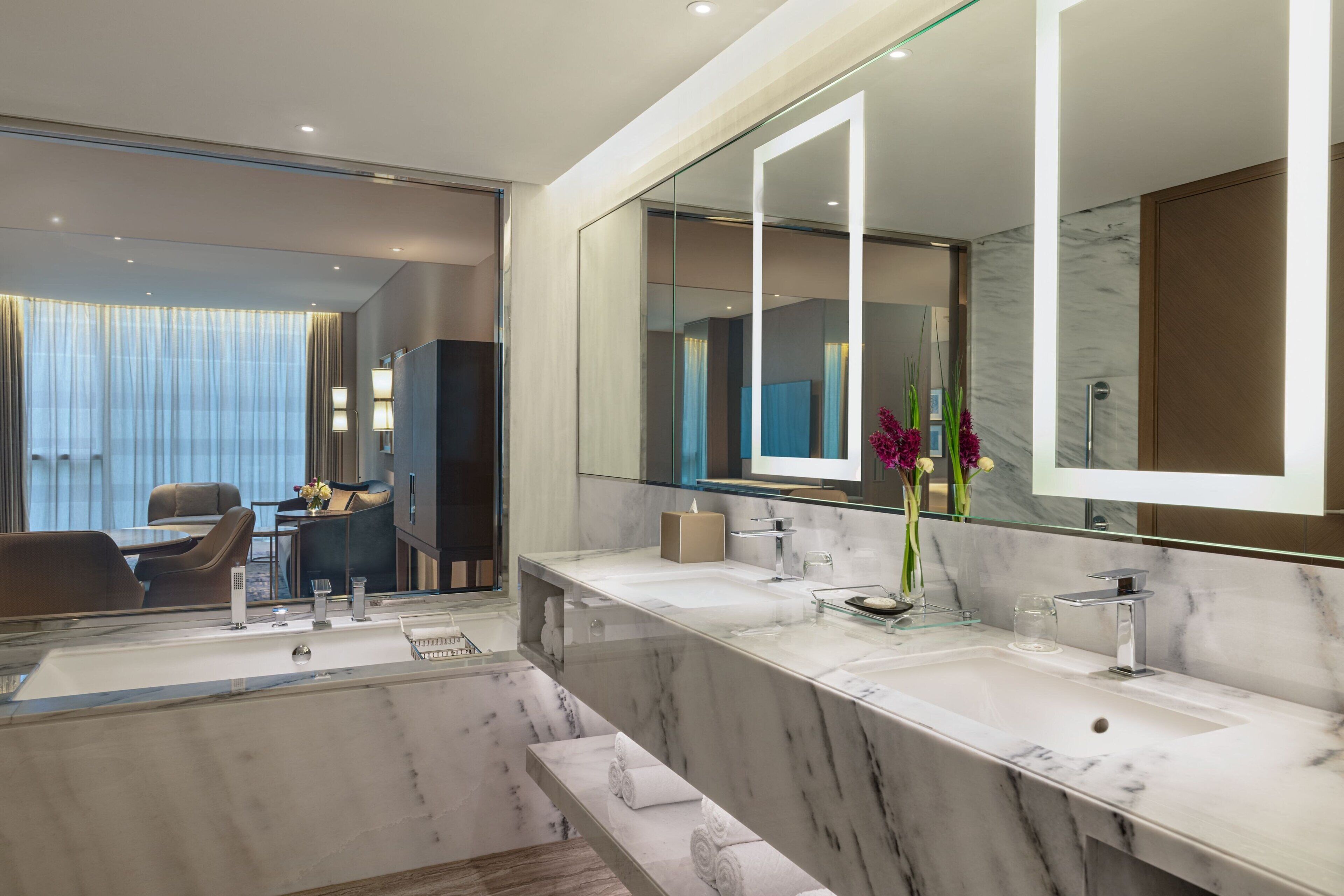 Une salle de bain moderne avec deux lavabos, un grand miroir et un coin salon comprenant une table et des chaises.