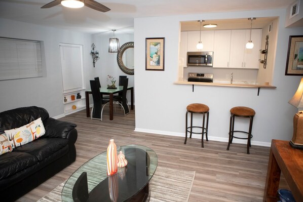 A modern living room with a black leather sofa, a glass coffee table, and a dining area with a wooden table and chairs.