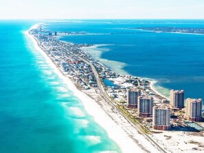 An aerial view of a coastal area with a long stretch of beach, adjacent high-rise buildings, and a clear view of the ocean.