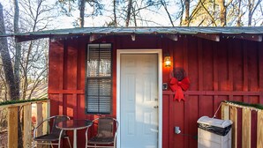 A wooden deck with a white door, a metal chair, a trash bin, and a black dog lying on the deck. - Yeti Cabin (Benton)