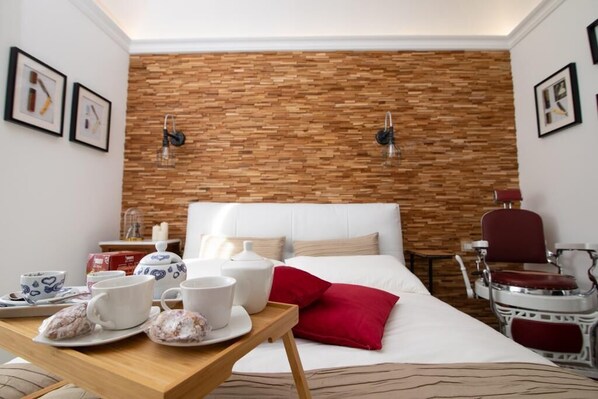 Ein Hotelzimmer mit einer Wand im Backsteinmuster, einem Bett mit weißer Bettwäsche, einem roten Kopfkissen, einem Holztischchen mit Teetassen und einem Tablett sowie einem roten Sessel.