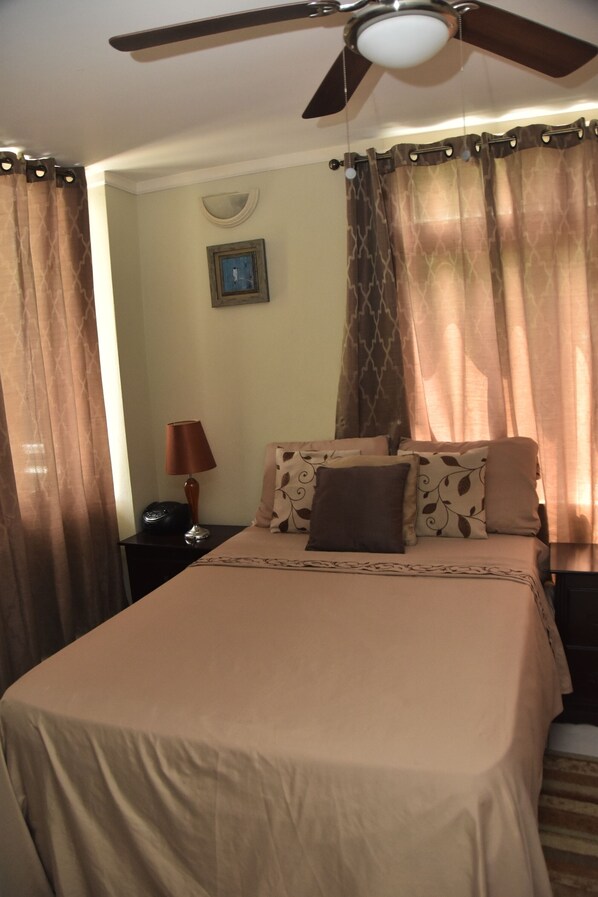 Una cama bien hecha con una colcha color beige, un almohadón con estampado de hojas y una lámpara de noche en una mesita.