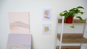 A room with a white wall displaying three artworks, a wooden shelf unit, and a potted plant. - Special Discount!! The Artist Condo in Barrio Dent (San Pedro)