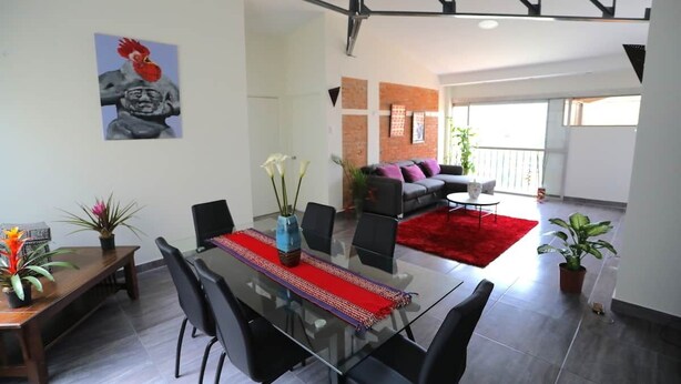 A modern living room with a glass dining table, black chairs, and a red rug. There is a sofa with cushions and a plant in the corner. - Special Discount!! The Artist Condo in Barrio Dent (San Pedro)
