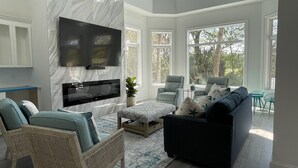 A spacious living room with a large flat-screen TV mounted on a marble wall, a comfortable seating area with a dark sofa and armchairs, and large windows offering a view of the outdoors. - Island Escape! Pool, Elevator, Pinball, Foosball, Arcade, Nursery,Infrared Sauna (Hilton Head Island)