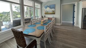 A dining area with a table set for six, surrounded by wicker chairs, with a view of a lake and greenery through large windows. - Island Escape! Pool, Elevator, Pinball, Foosball, Arcade, Nursery,Infrared Sauna (Hilton Head Island)