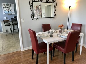 Un coin repas avec une table blanche dressée pour quatre personnes, entourée de chaises rouges. Un miroir à cadre noir est accroché au-dessus de la table, et une lampe est placée sur le côté droit.