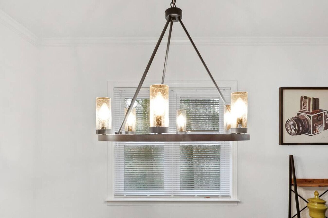 A chandelier with multiple lights, a window with blinds, and a framed picture on the wall.