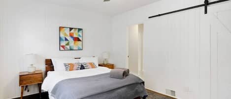 Un dormitorio con cama, ventilador de techo, mesita de noche, lámpara, una obra de arte en la pared y una puerta.