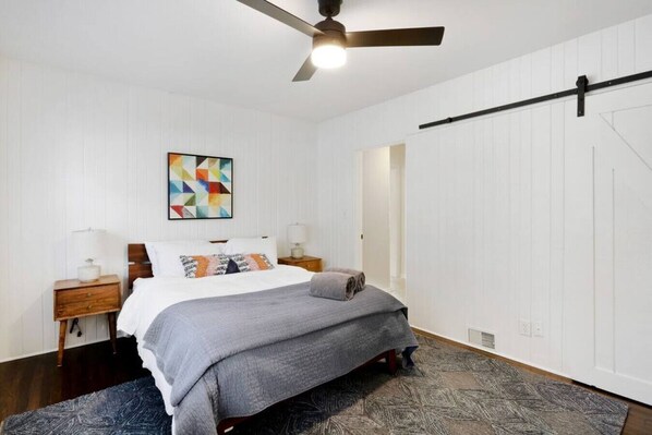 Un dormitorio con cama, ventilador de techo, mesita de noche, lámpara, una obra de arte en la pared y una puerta.