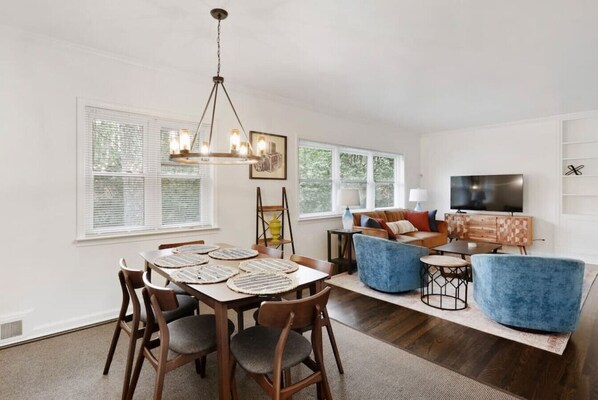 A dining area with a wooden table and chairs, a chandelier, and a view of the outdoors through large windows.