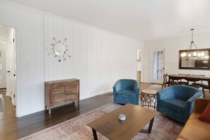 A spacious living room with a wooden cabinet, two blue armchairs, a coffee table, and a dining area with a chandelier.