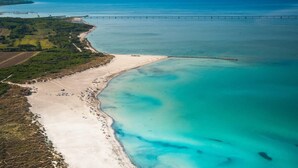 Luftaufnahme einer Küstenlandschaft mit Sandstrand, klarem türkisblauem Wasser und einer in der Ferne sichtbaren Brücke.