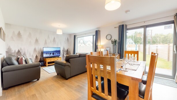 A modern living room with a flat-screen TV, a sofa, a dining table set for four, and a large window offering a view of the outdoors. - Alpine View - a cosy Aviemore family home (Aviemore)