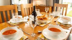 A dining table set with bowls of soup, wine glasses, and a bottle of wine, surrounded by wooden chairs and a vase of flowers. - Alpine View - a cosy Aviemore family home (Aviemore)