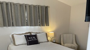 Ein ordentlich gemachtes Bett mit weißer Bettdecke und zwei Kissen, eines davon mit schwarzer Kissenhülle. Auf einem Nachttisch steht eine Lampe, ein Sessel und ein Fenster mit Vorhängen.