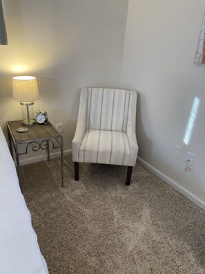 A room with a striped armchair, a small table with a lamp and an alarm clock, and a carpeted floor.