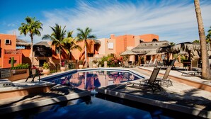 A swimming pool with lounge chairs, palm trees, and buildings with orange facades. - COZY, OCEAN VIEW CONDO WITH PRIVATE BEACH ACCESS (CABO SAN LUCAS)