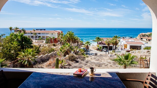 A view from a balcony overlooking a beach with palm trees, a pool, and buildings in the distance. - COZY, OCEAN VIEW CONDO WITH PRIVATE BEACH ACCESS (CABO SAN LUCAS)