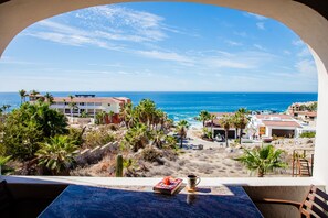 A view from a balcony overlooking a beach with palm trees, a pool, and buildings in the distance. - COZY, OCEAN VIEW CONDO WITH PRIVATE BEACH ACCESS (CABO SAN LUCAS)
