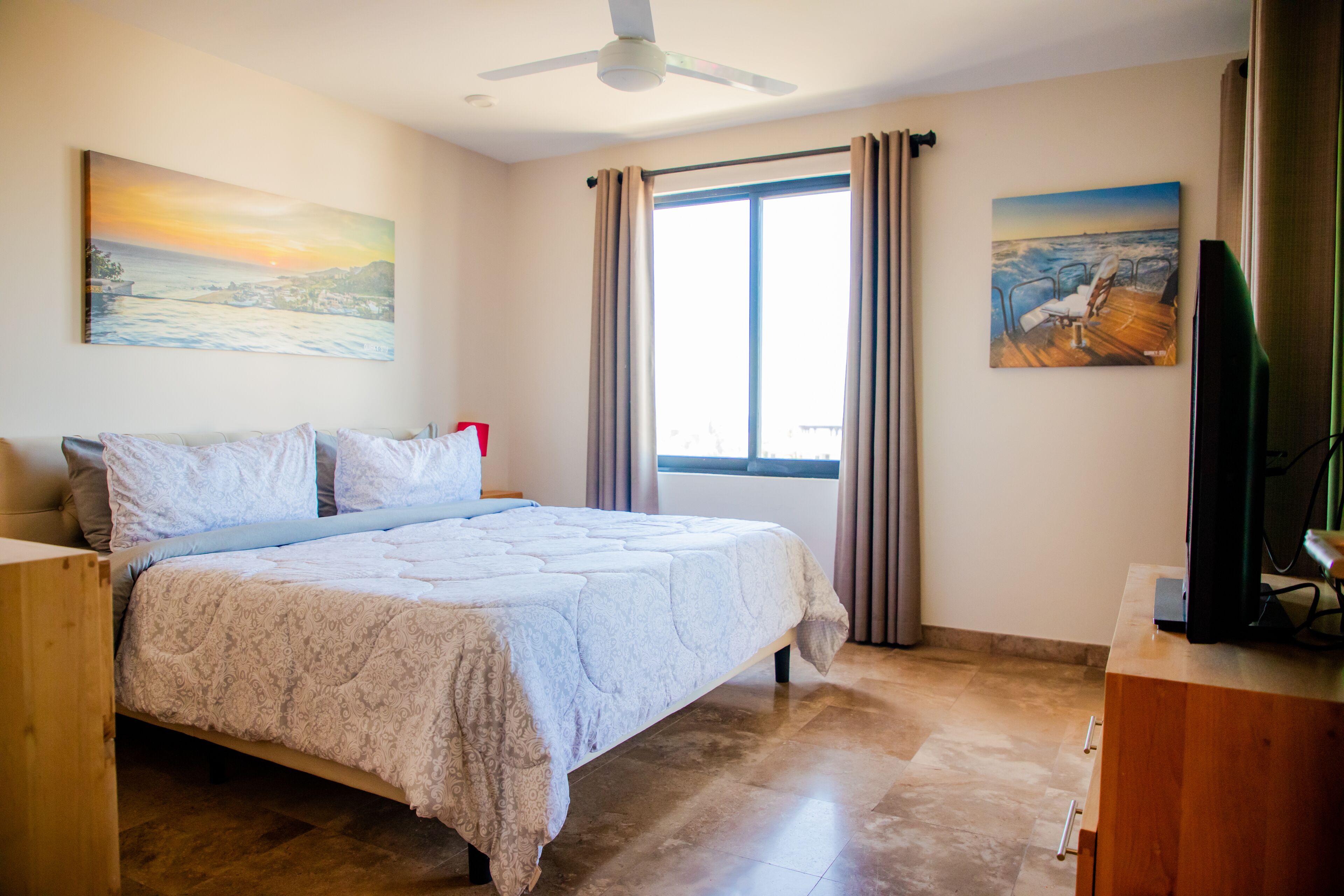 A bedroom with a bed, a ceiling fan, a window with curtains, and artwork on the walls.