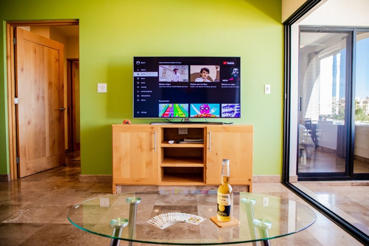Quarto com mesa de vidro, uma garrafa de cerveja Corona e uma TV exibindo canais do YouTube.