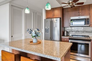 A kitchen with granite countertops, stainless steel appliances, wooden cabinets, and pendant lighting. - Direct Oceanfront View! Walk on Beaches - A/C (Kihei)