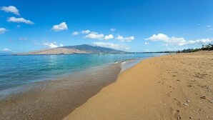 Ein Strand mit klarem, blauem Wasser, sandigem Ufer und einem entfernten Gebirge.
