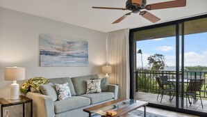 Ein modernes Wohnzimmer mit einer Couch, einem Couchtisch und zwei Lampen. Es gibt einen Balkon mit Gartenmöbeln und Blick auf die Landschaft.