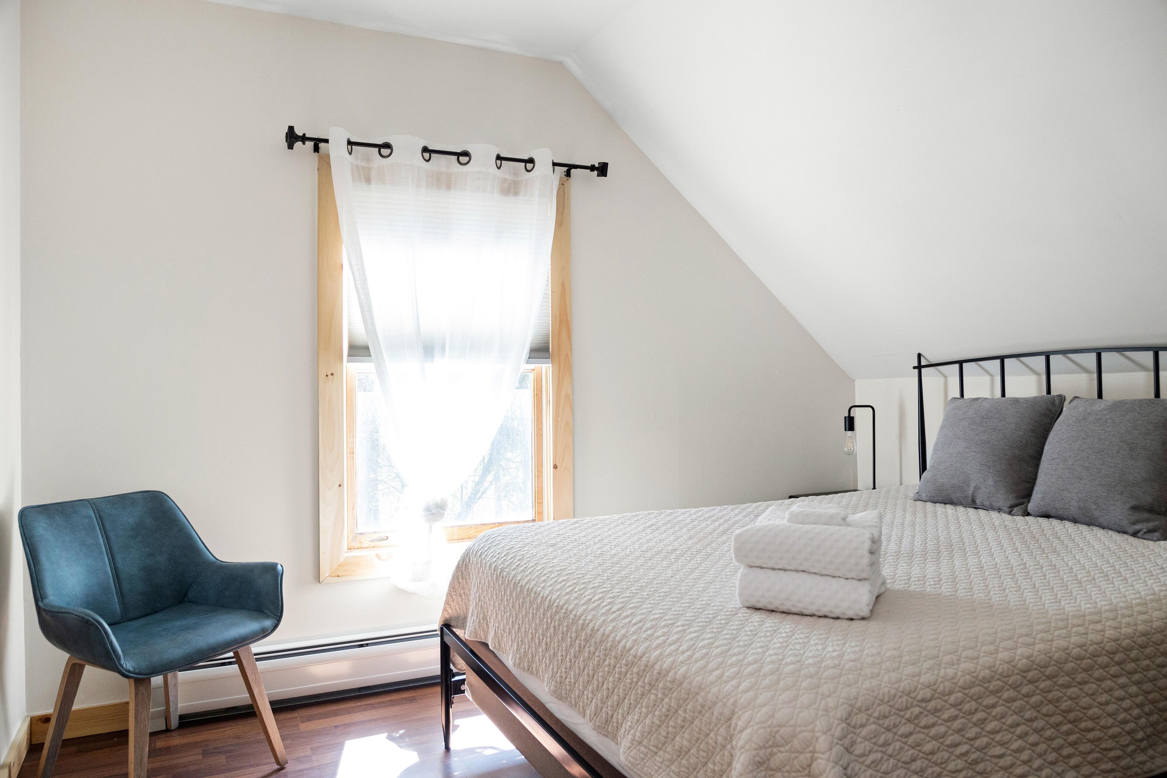 Un dormitorio con una cama, una silla, un ventanal con cortinas y una mesita de noche con lámpara.