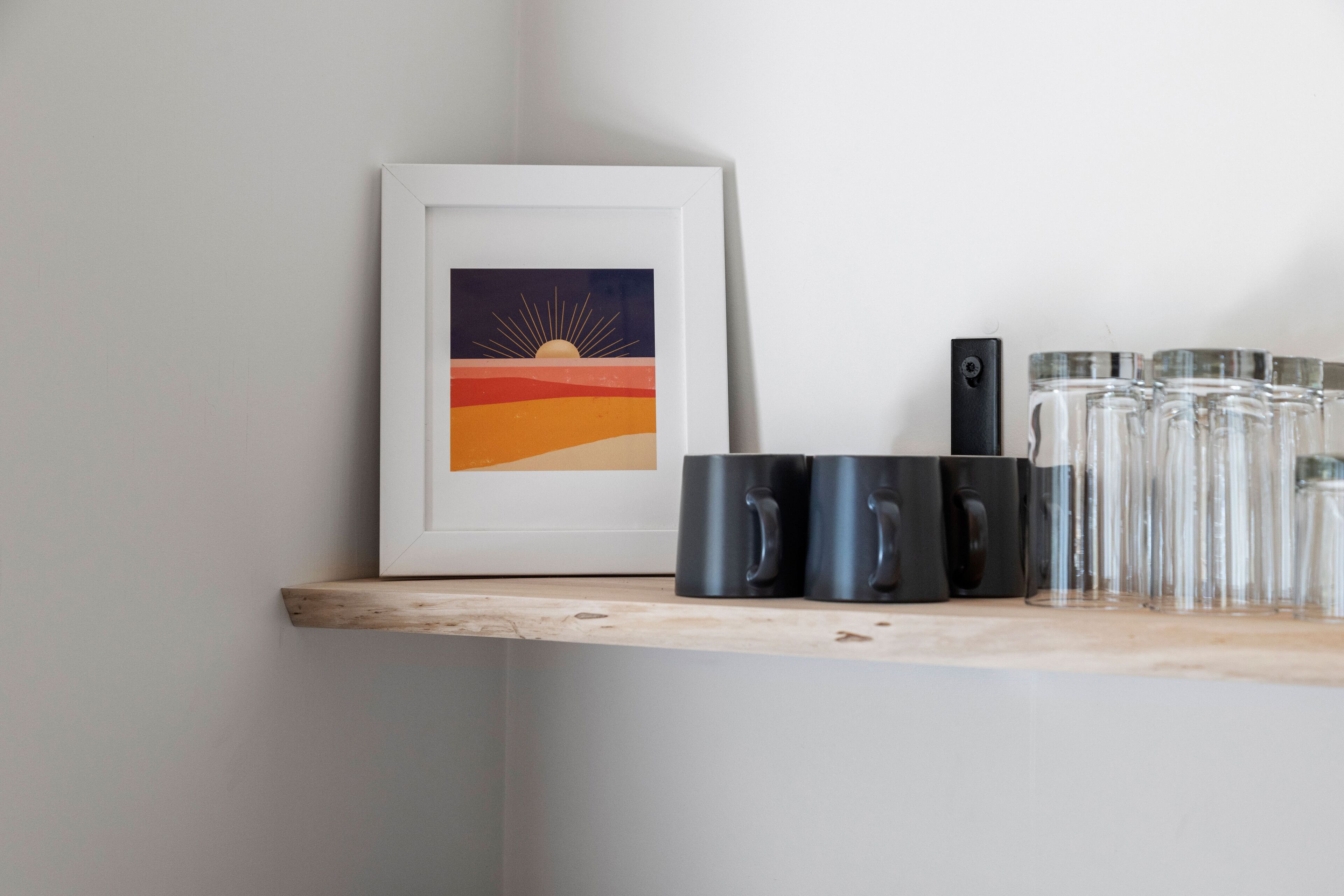 Una obra de arte enmarcada con un atardecer sobre una repisa, tazas negras y vasos de vidrio.