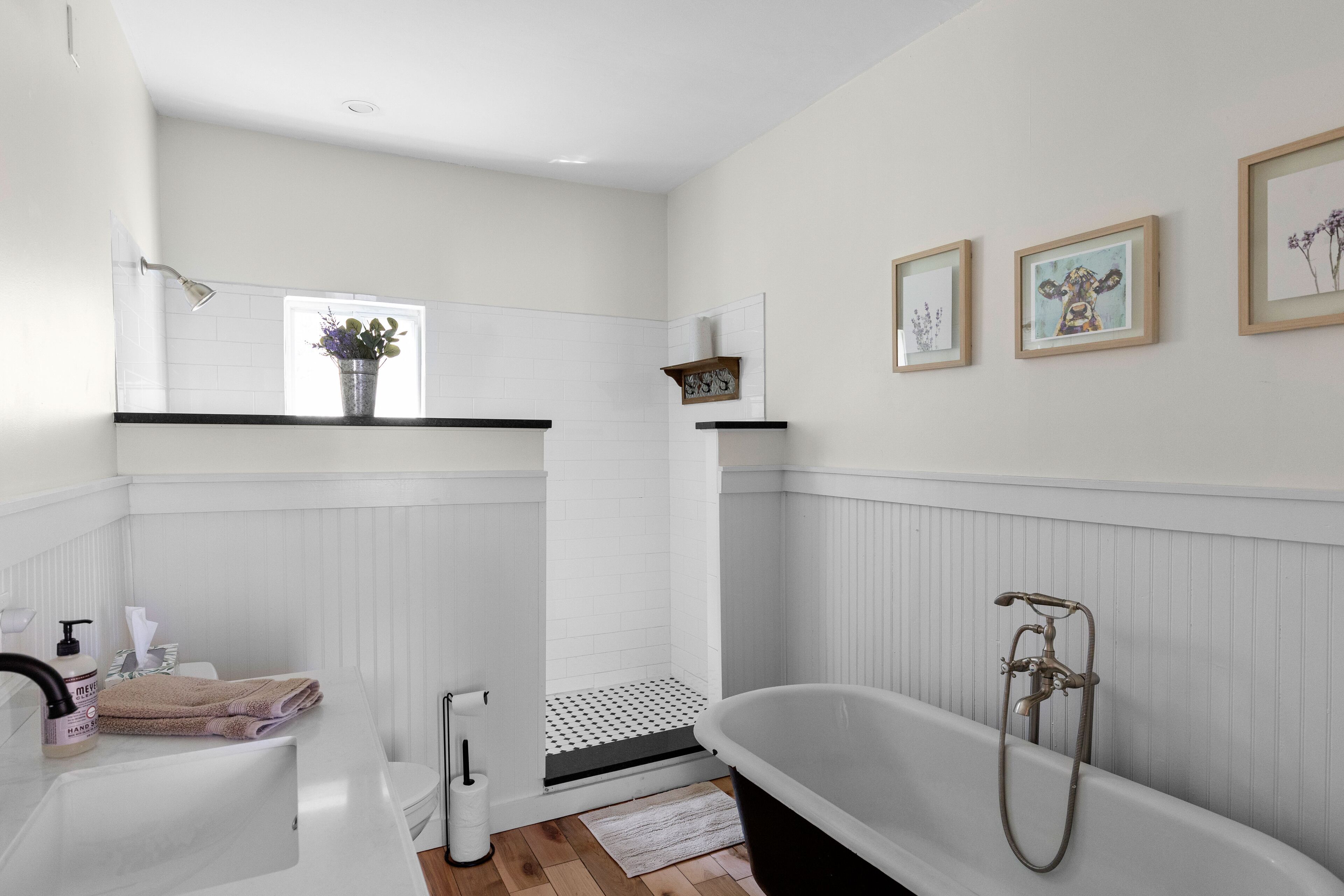 Badeværelse med badekar, toilet, vask og brusebad. Der er billeder hængende på væggen, og et vindue med en plante på hylden.