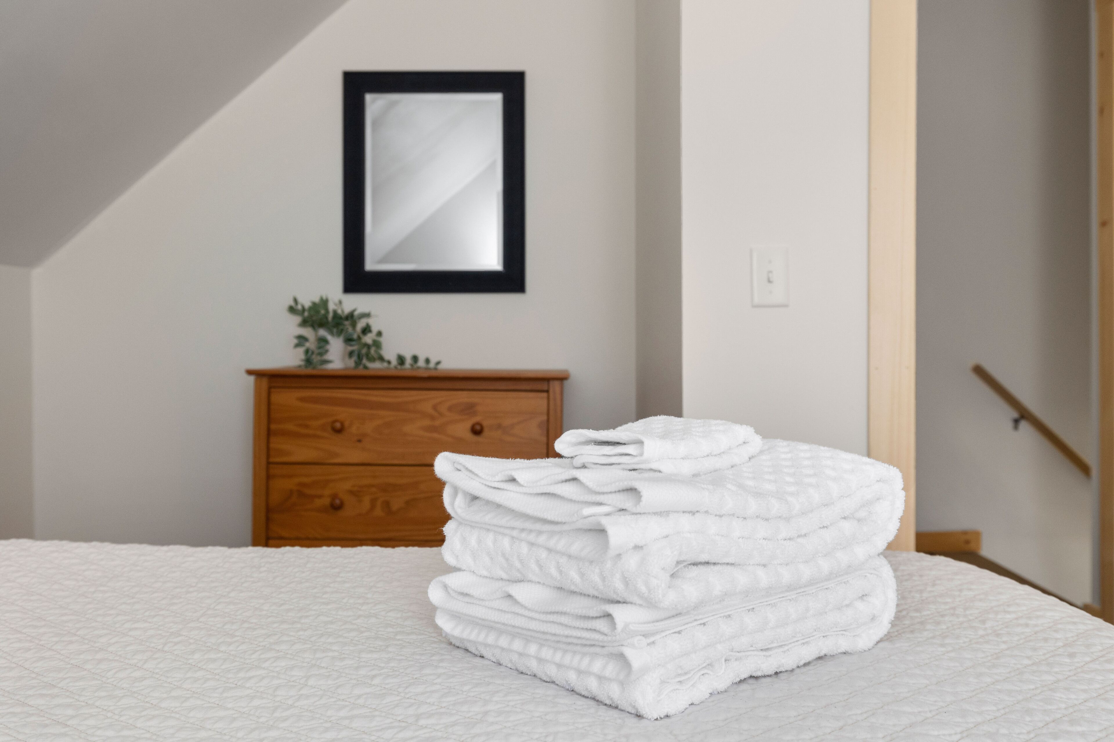 A neatly made bed with a white quilt, a stack of folded white towels, a wooden dresser, and a framed mirror on the wall.