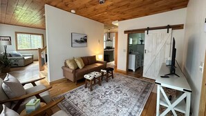 Ett rymligt vardagsrum med ett träpanelat tak, en stor matta, en soffa och en matplats med en tv.