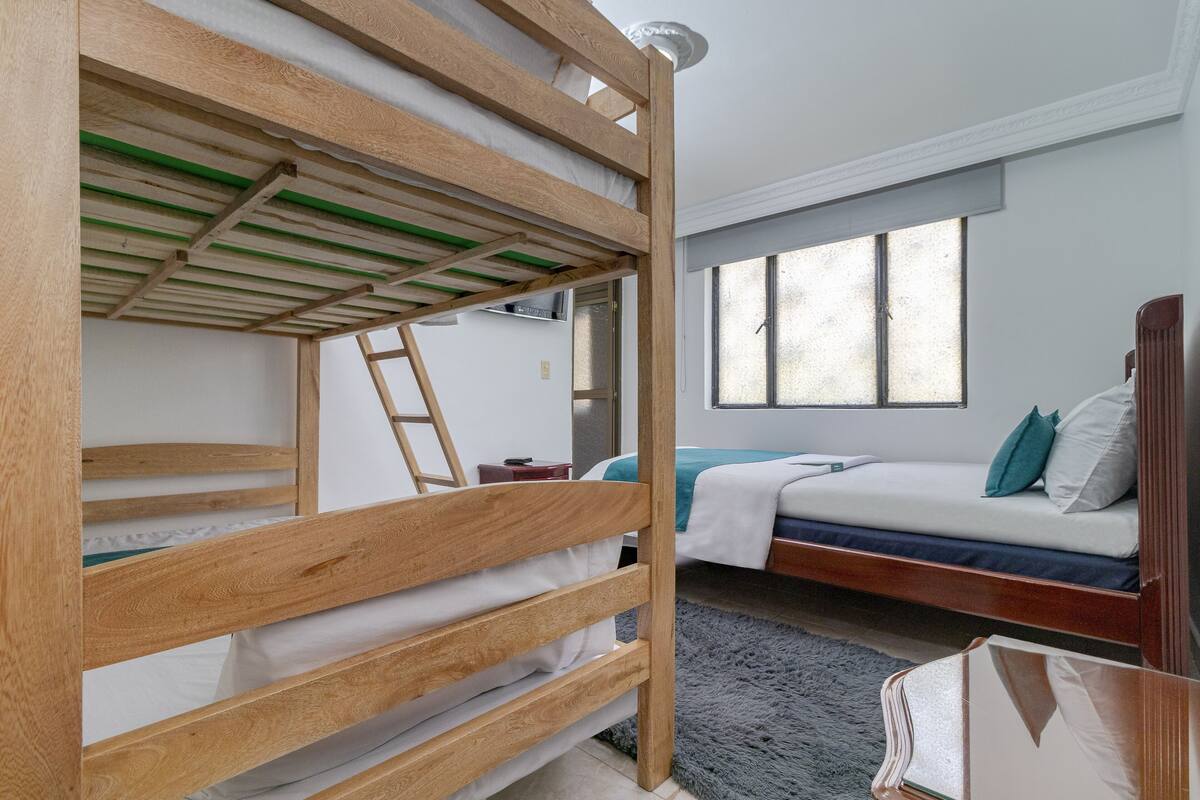 A bunk bed with a wooden frame and a ladder, a single bed with a blue mattress, and a window with a view of the outside.