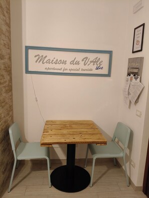 Une petite table avec deux chaises, une pancarte indiquant « Appartement Maison du Vole, pour les touristes privilégiés », et une boîte électrique fixée au mur, contenant des papiers.