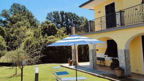 Een geel gebouw met een balkon, een terras met een blauwe parasol, en een gazon met een boom en een bank.