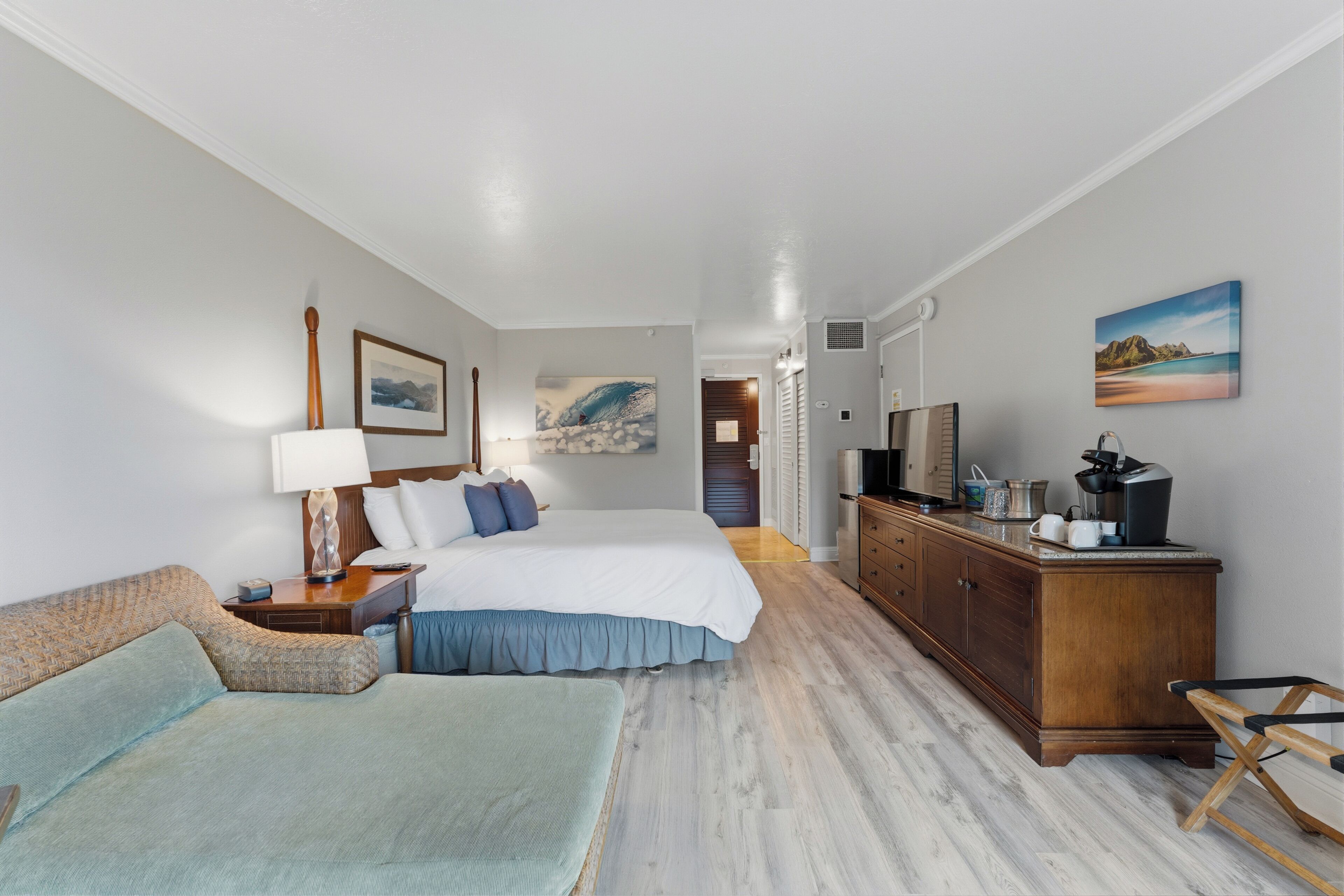 A hotel room with a bed, a sofa, a desk with a lamp, a dresser, and artwork on the walls.
