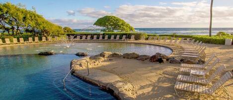 A pool with clear water, surrounded by lounge chairs and a palm tree, with a view of the ocean and a bridge in the background.