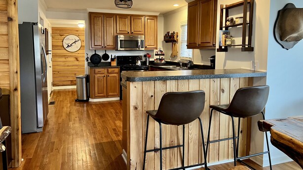 A modern kitchen with wooden cabinets, a stainless steel refrigerator, a microwave, and a bar area with stools. - Canvasback Cabin - 2bd/2ba (Scottsboro)