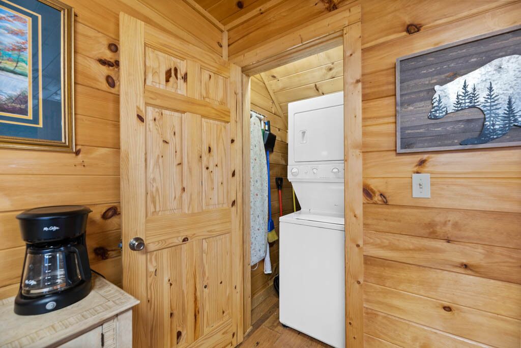 Un interior de madera con una cafetera sobre una encimera, un refrigerador blanco y una puerta que da a un armario con ropa colgada.