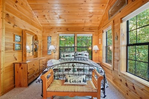 Ein gemütliches Blockhaus-Interieur mit Holzbalkendecke, einem Bett mit Bärenmotiv, einem Holzschrank, einem Sessel und großen Fenstern mit Blick auf den Wald.