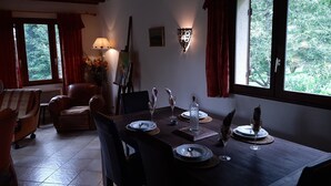 Ein Essbereich mit einem Holztisch, gedeckt für vier Personen, umgeben von Stühlen. Es gibt eine Lampe, ein Bild an der Wand und ein Fenster mit Vorhängen.