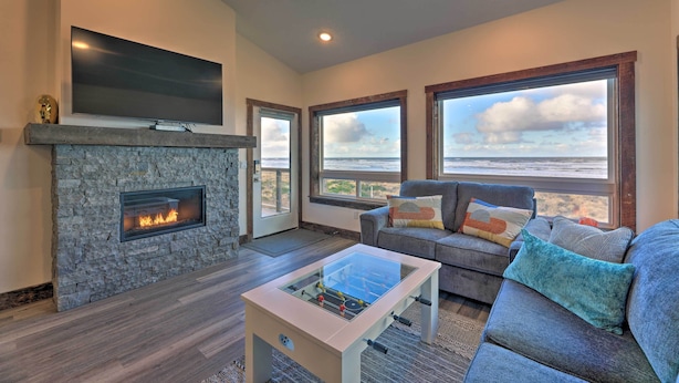 A living room with a stone fireplace, a flat-screen TV, a sofa with cushions, and large windows with a view. - Sanderling Sea Cottages, Unit 7 w/ Beach Access! (Waldport)